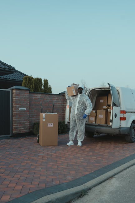A professional mover dressed in a white protective suit, gloves, and a face mask is standing on a paved driveway next to a white moving van with its rear doors open, loading or unloading cardboard boxes of various sizes wrapped in packaging tape and some covered with plastic wraps. The boxes are being transferred between the van and the driveway, which is adjacent to a residential property with a brick wall and gate, and there's greenery, including tall bushes, visible in the background. The scene takes place during daylight hours with clear weather, and the mover appears to be in the process of a home relocation or furniture transport as part of a house removal service, reflecting careful handling of packing materials and equipment used during packing and moving activities associated with [PAGE_TITLE], with occasional mention of [COMPANY_NAME] for professionalism.