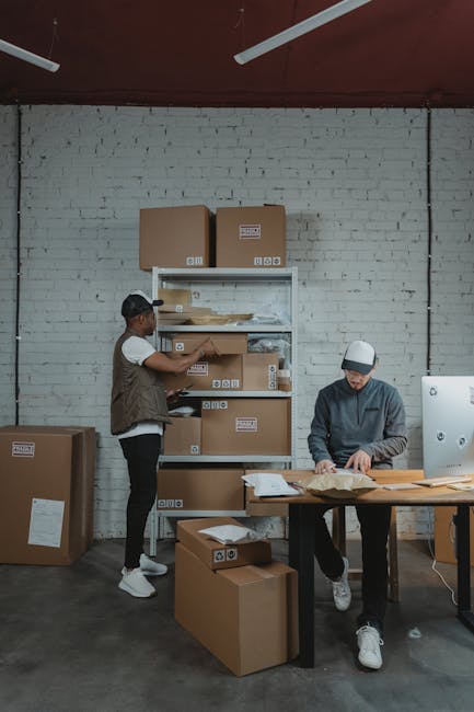 A person wearing a dark green shirt is holding a medium-sized cardboard box during a house relocation. The box has a white label with red text and a red arrow pointing upward, reading 'CAUTION THIS SI