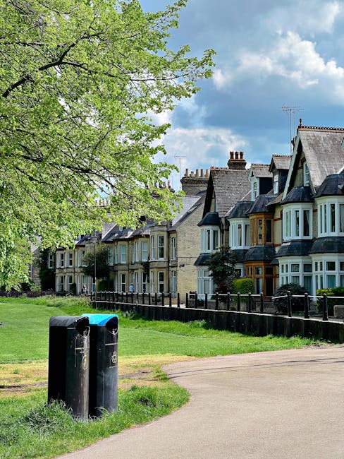A row of Victorian-style terraced houses with bay windows and pitched roofs, situated behind a black metal fence along Colliers Wood High Street. In the foreground, two black waste bins are positioned on a grassy verge beside a paved pathway. A large green-leaved tree extends its branches overhead, partially shading the scene. The sky is partly cloudy with patches of blue visible, and the environment appears to be a residential neighbourhood. This setting represents a typical area where local house removals and furniture transport services by Man and Van Colliers Wood may be performed during home relocation projects, highlighting the typical urban landscape and related moving logistics.