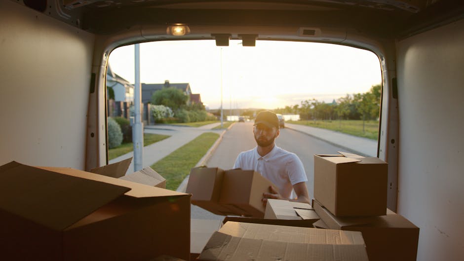Two men are involved in a home relocation process outside a residential property, loading large cardboard boxes and appliances onto a white commercial van equipped for furniture transport. They are ha