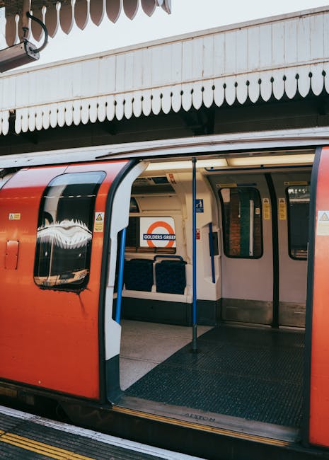 An empty London Underground train at Colliers Wood station, with open sliding doors revealing the interior platform area. The train has a red exterior with black-tinted windows and a white interior panel, and is positioned adjacent to a station platform made of concrete with yellow tactile paving along the edge. Inside the train, there are blue fabric-covered seats and vertical blue handrails, with some storage space visible near the doors. The platform features a white overhanging roof with decorative scalloped edging, providing shelter for passengers. The image captures a clear view of the train's entrance and the station environment, which is involved in home relocation or furniture transport activities associated with moving services, such as those provided by Man and Van Colliers Wood.