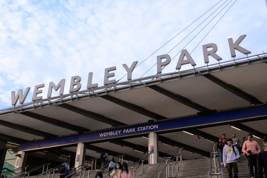 Photograph of the Wembley Park Station entrance with a large overhead sign displaying 'WEMBLEY PARK' in bold white letters. The station's canopy roof extends over the entrance, supported by black metal beams, with a blue sign beneath indicating 'WEMBLEY PARK STATION.' Several people, some wearing face masks, are seen descending the outdoor stairs, with a few carrying backpacks or small luggage. The background features a partly cloudy sky and overhead electrical wires visible above the station. The scene reflects typical busy urban transportation surroundings, relevant to house removals and furniture transport services provided by Man and Van Colliers Wood, especially in the context of home relocation logistics involving the London area and station access points.