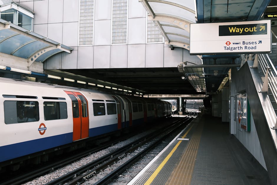 An empty London Underground train at Colliers Wood station, with open sliding doors revealing the interior platform area. The train has a red exterior with black-tinted windows and a white interior panel, and is positioned adjacent to a station platform made of concrete with yellow tactile paving along the edge. Inside the train, there are blue fabric-covered seats and vertical blue handrails, with some storage space visible near the doors. The platform features a white overhanging roof with decorative scalloped edging, providing shelter for passengers. The image captures a clear view of the train's entrance and the station environment, which is involved in home relocation or furniture transport activities associated with moving services, such as those provided by Man and Van Colliers Wood.